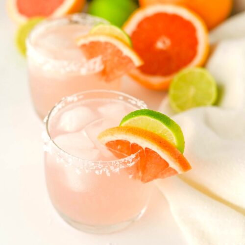 Two glasses containing pink liquid with salt on rims, fruit slices on rim, and white napkin, grapefruit, limes, and plant in background.