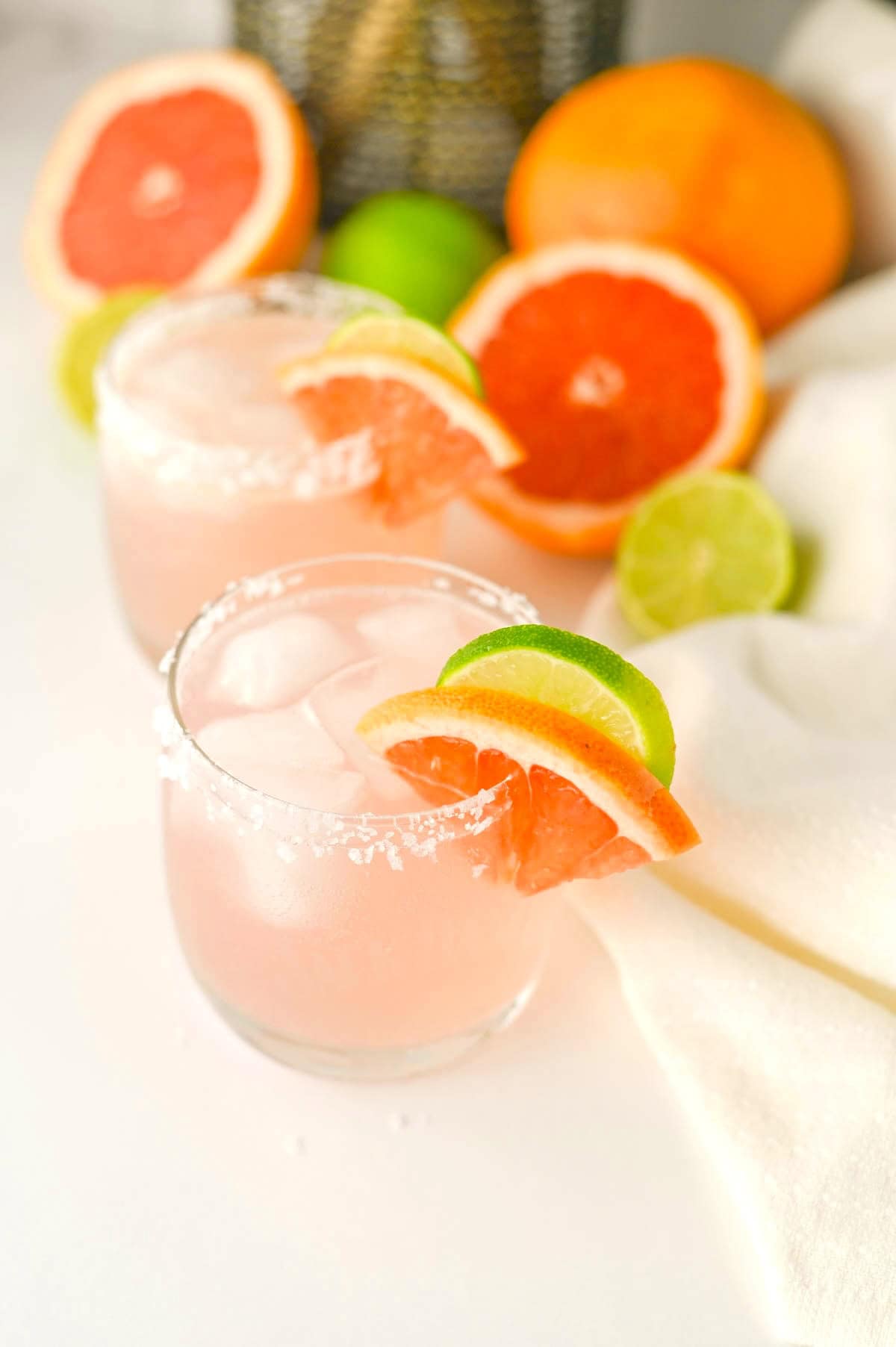 Two glasses containing pink liquid with salt on rims, fruit slices on rim, and white napkin, grapefruit, limes, and plant in background.