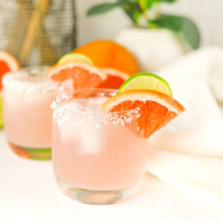 Two glasses containing pink liquid with salt on rims, fruit slices on rim, and white napkin, grapefruit, and plant in background.