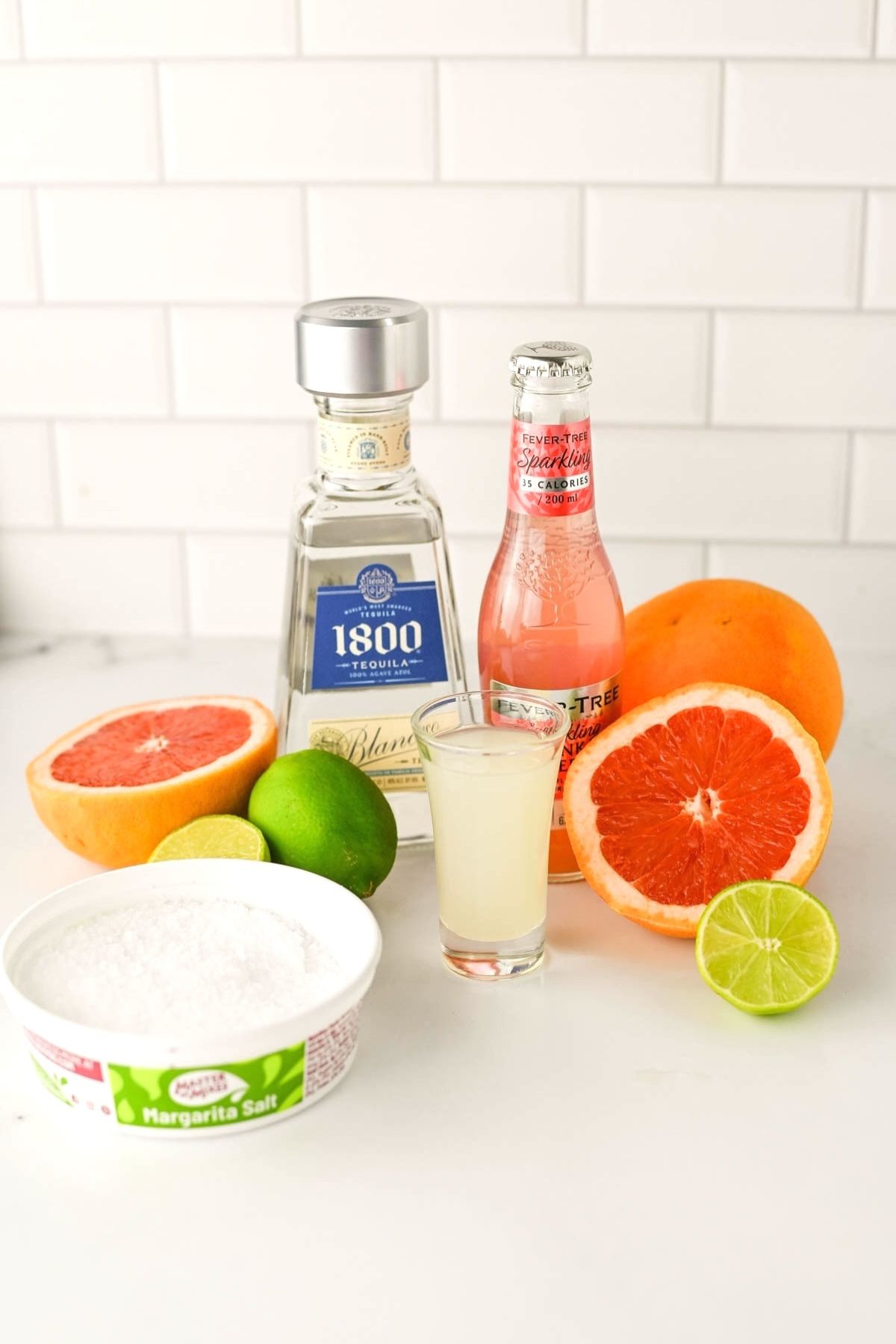 Bottle of tequila, bottle of pink liquid,  container of salt, lime juice, a grapefruit and a lime on a white counter.
