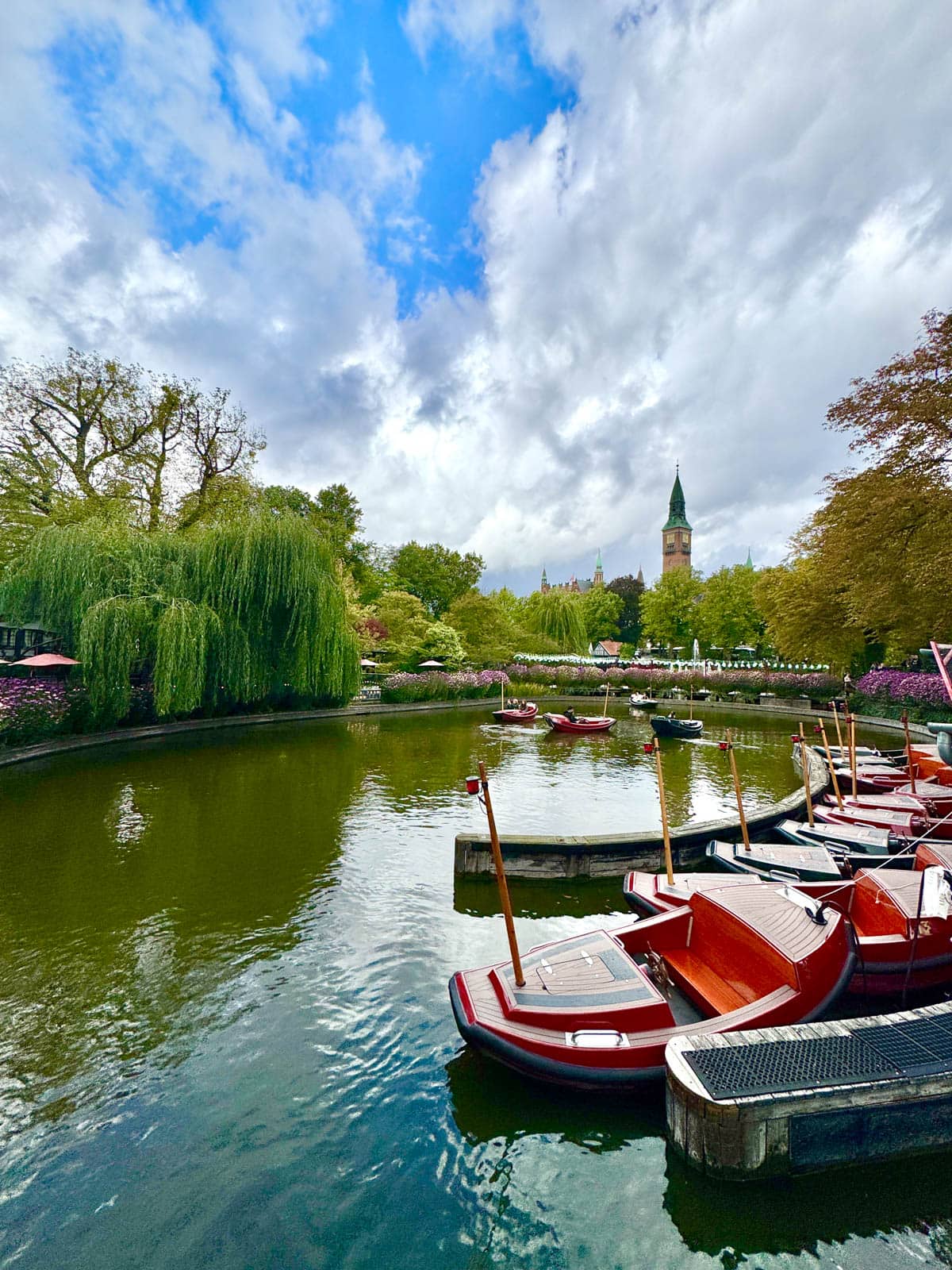 Pond with small Asian inspired boats with a lot of green trees and gardens and tall tower like structure in background against a blue sky.