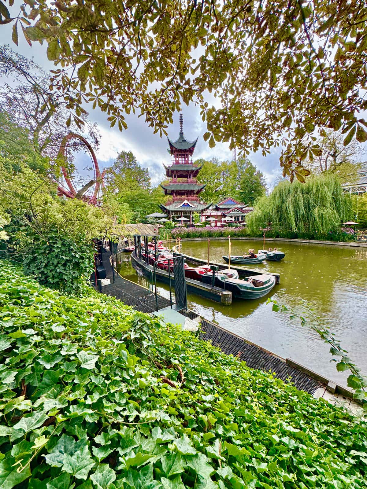 Chinese temple with pond and small boats in front with a lot of greenery all around and roller coaster off to the left.