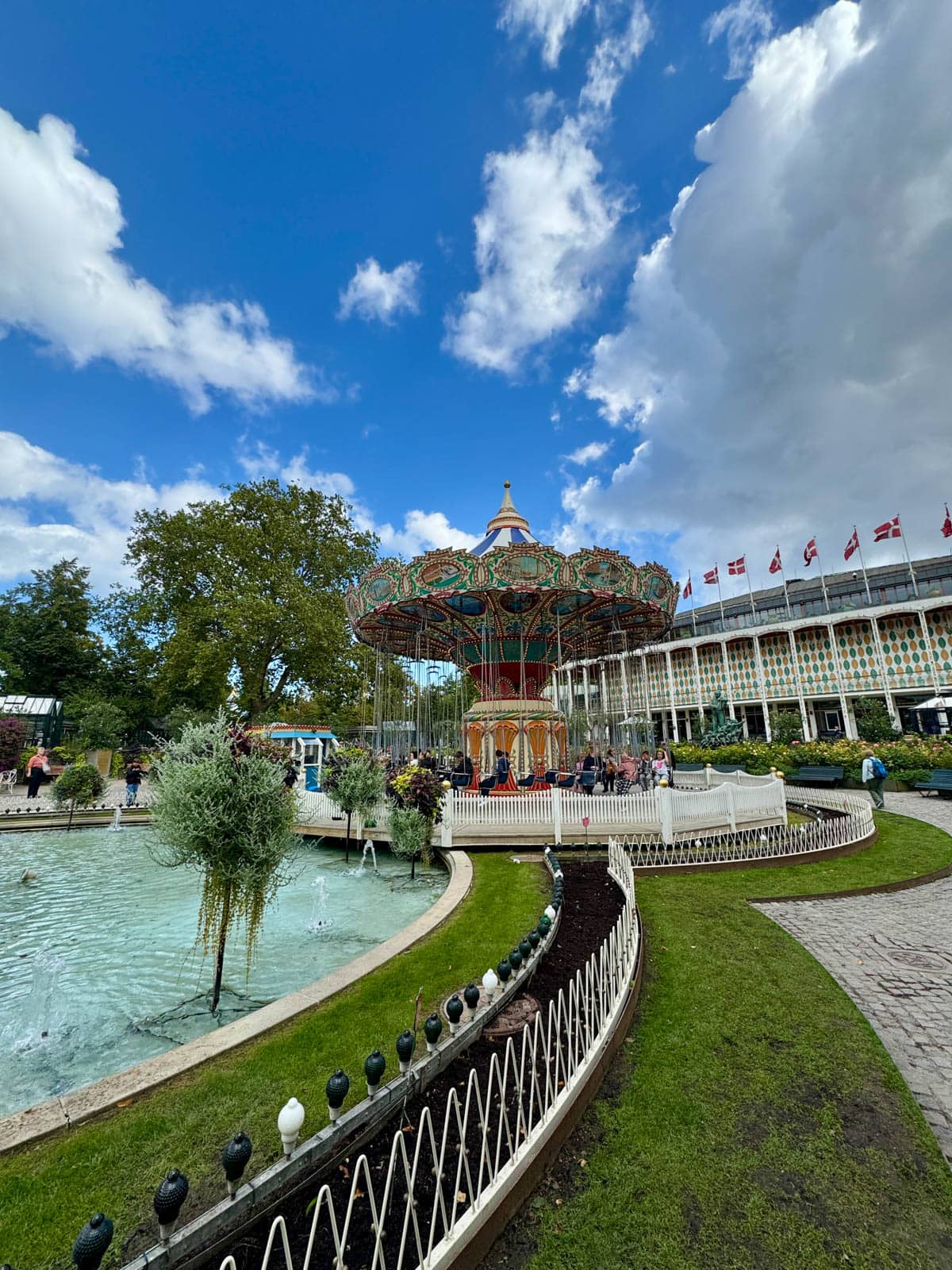 Merry-go-round against a blue cloudy sky next to a pond with a lot of greenery around.