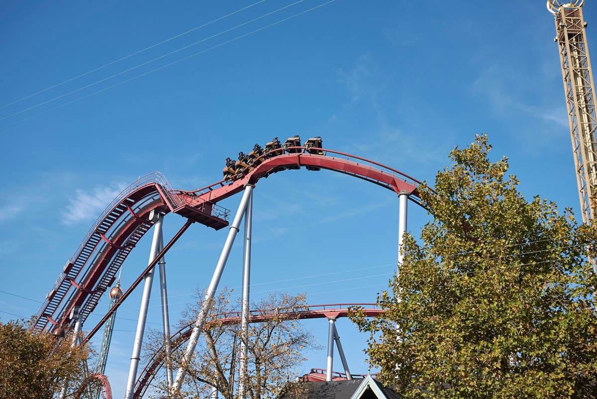 Old wooden roller coaster with cars at top against a blue sky with green tree to the side.