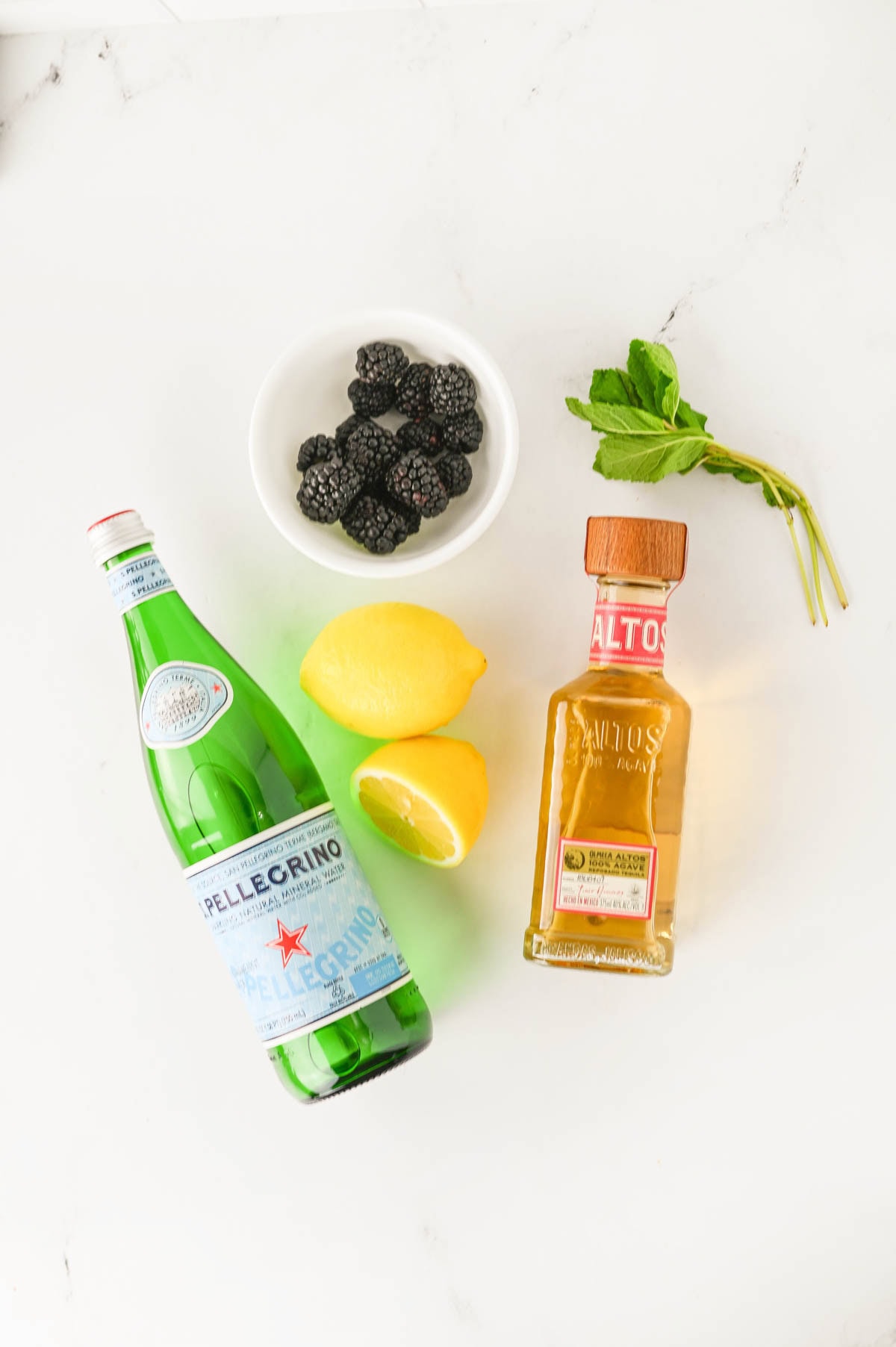 Bottle of soda water, lemons, white bowl of blackberries, bundle of mint, and bottle of tequila.