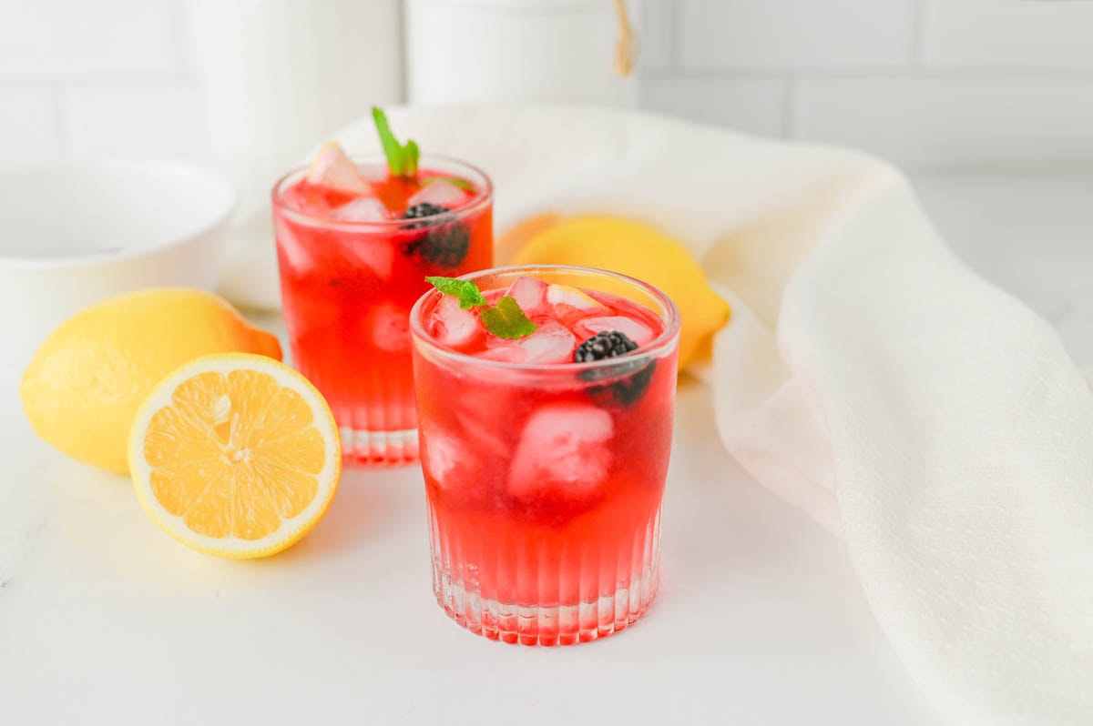 Purple cocktail in a rocks glass with ice, blackberry, and mint, with lemons on the sides laying on a white counter with kitchen background.