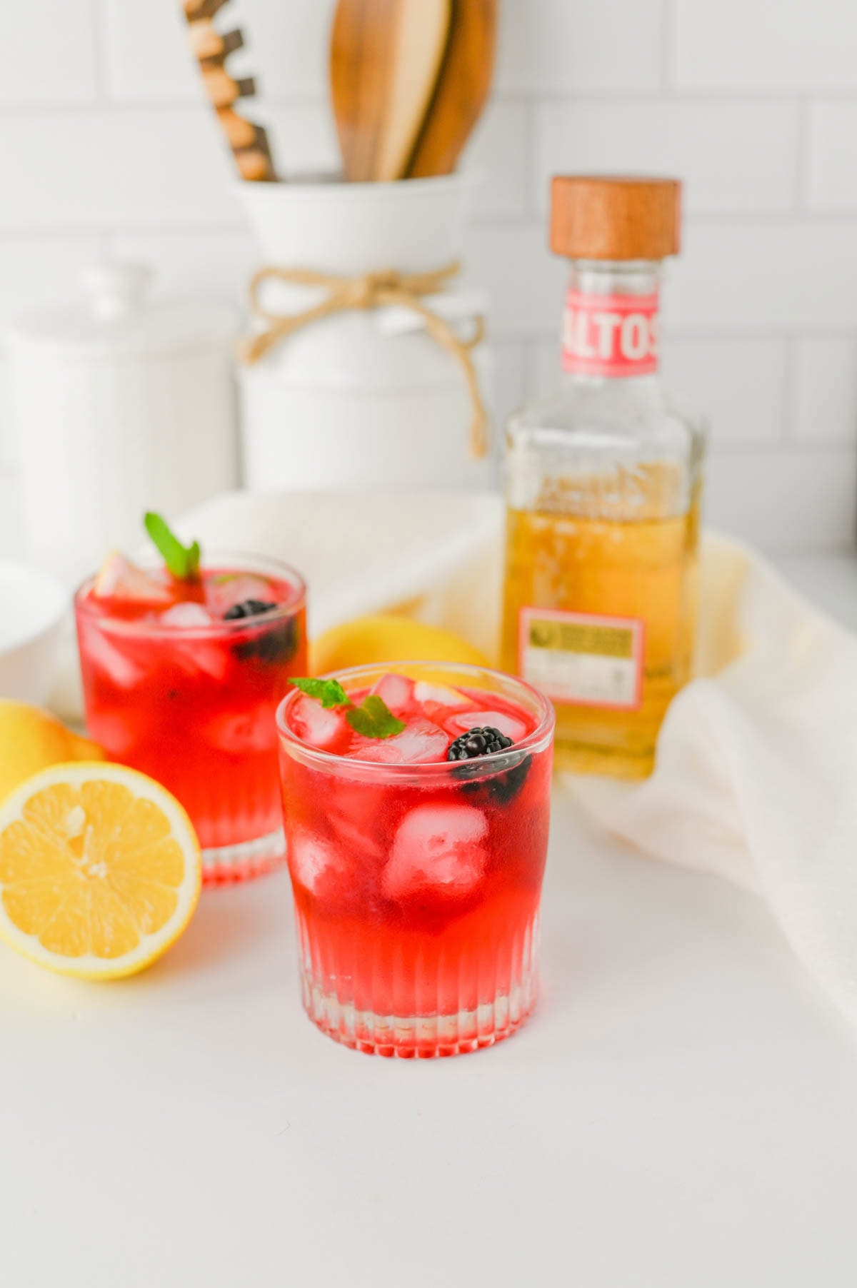 Purple cocktail in a rocks glass with ice, blackberry, and mint, with lemons on the sides laying on a white counter next to bottle of tequila with kitchen background.