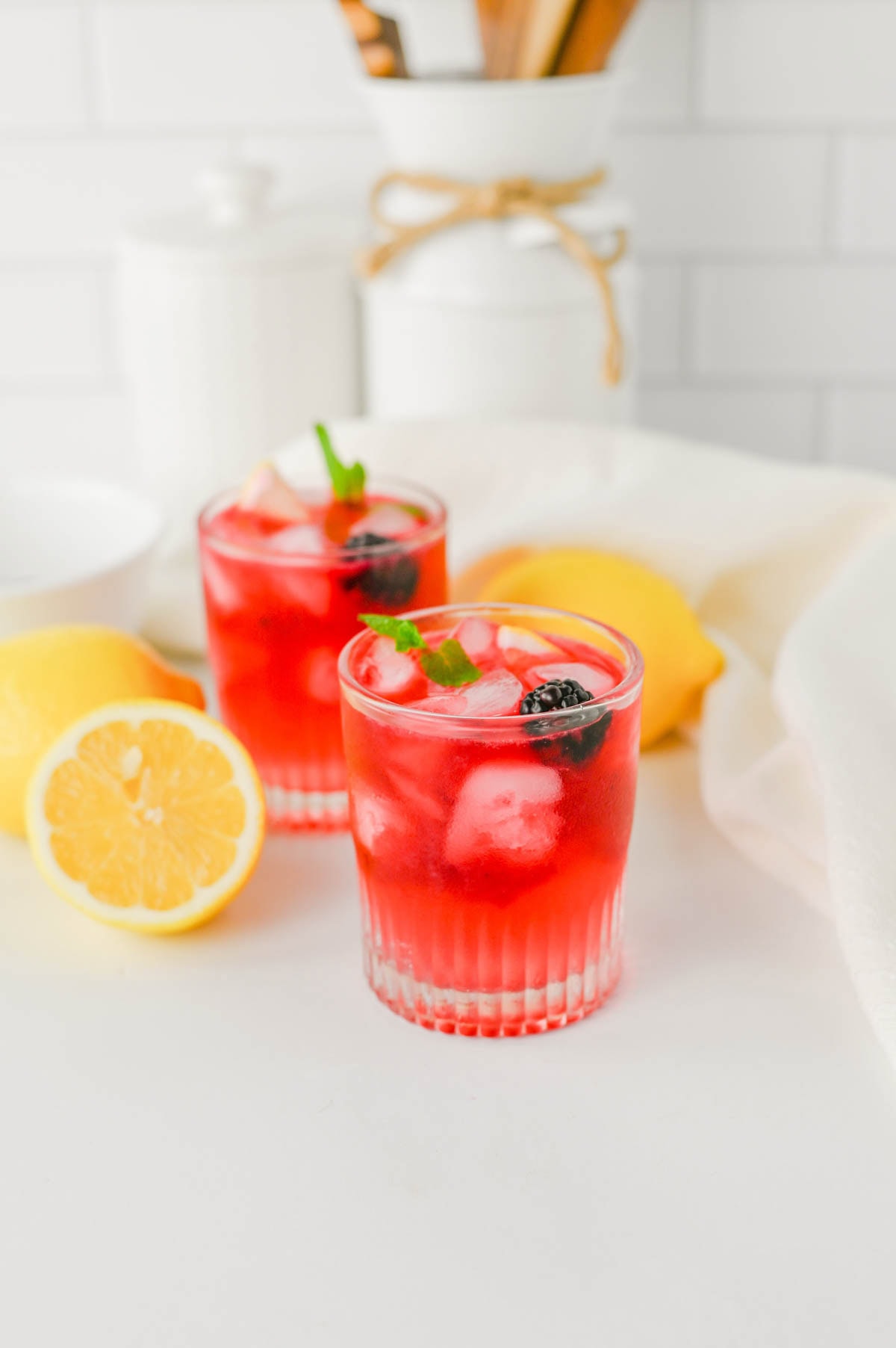 Purple cocktail in a rocks glass with ice, blackberry, and mint, with lemons on the sides laying on a white counter with kitchen background.