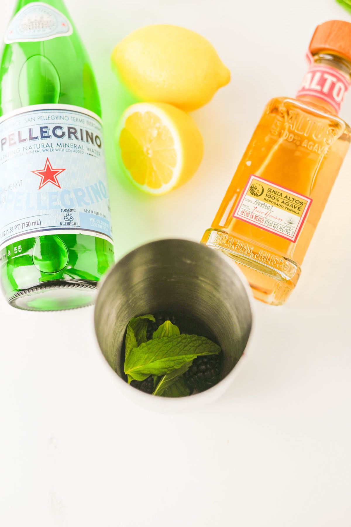 Bottle of soda water, lemons, bottle of tequila, and cocktail shaker with blackberries and mint inside.