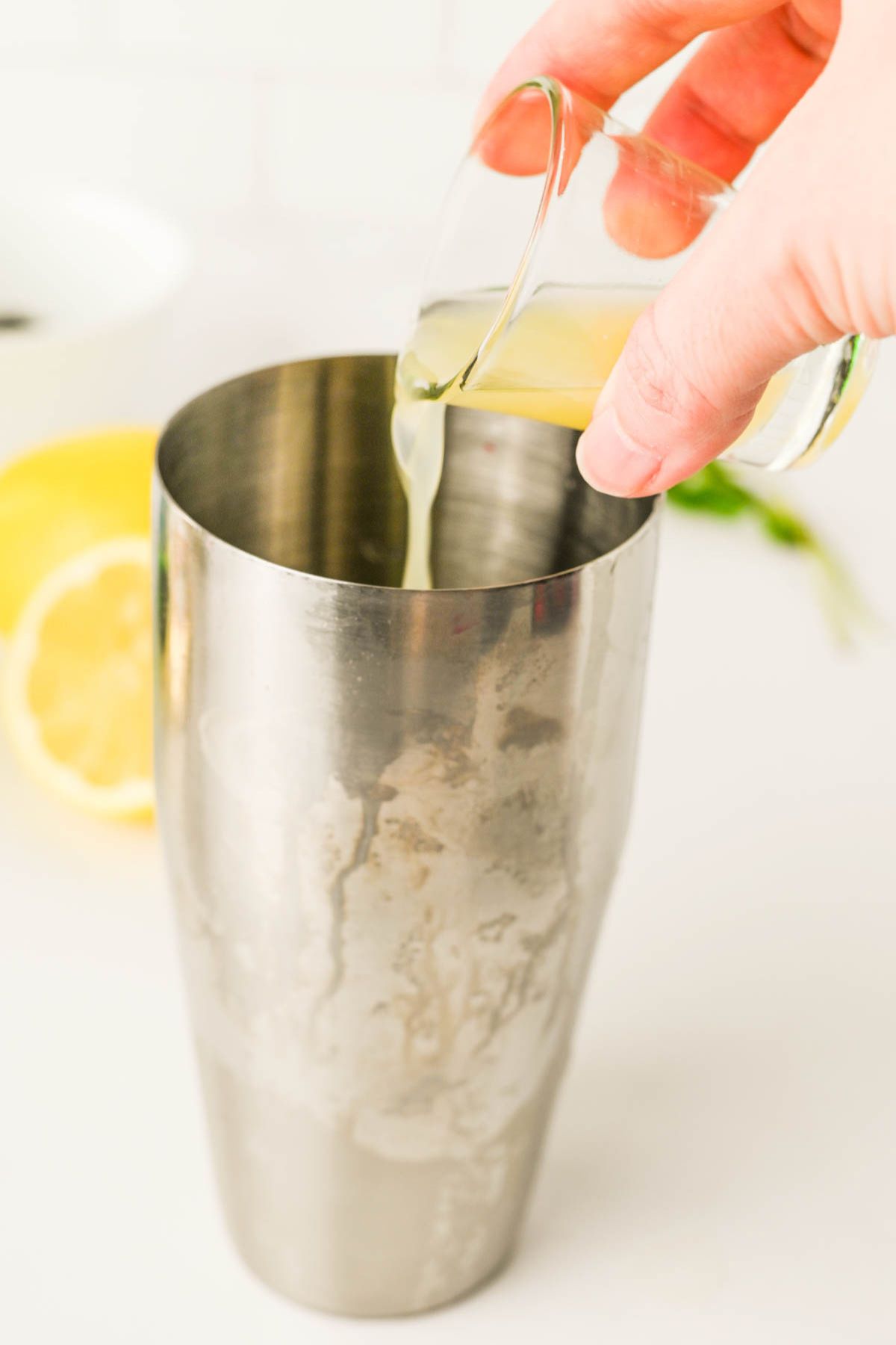 Stainless cocktail shaker with hand pouring shot of lemon juice in with lemons in background.