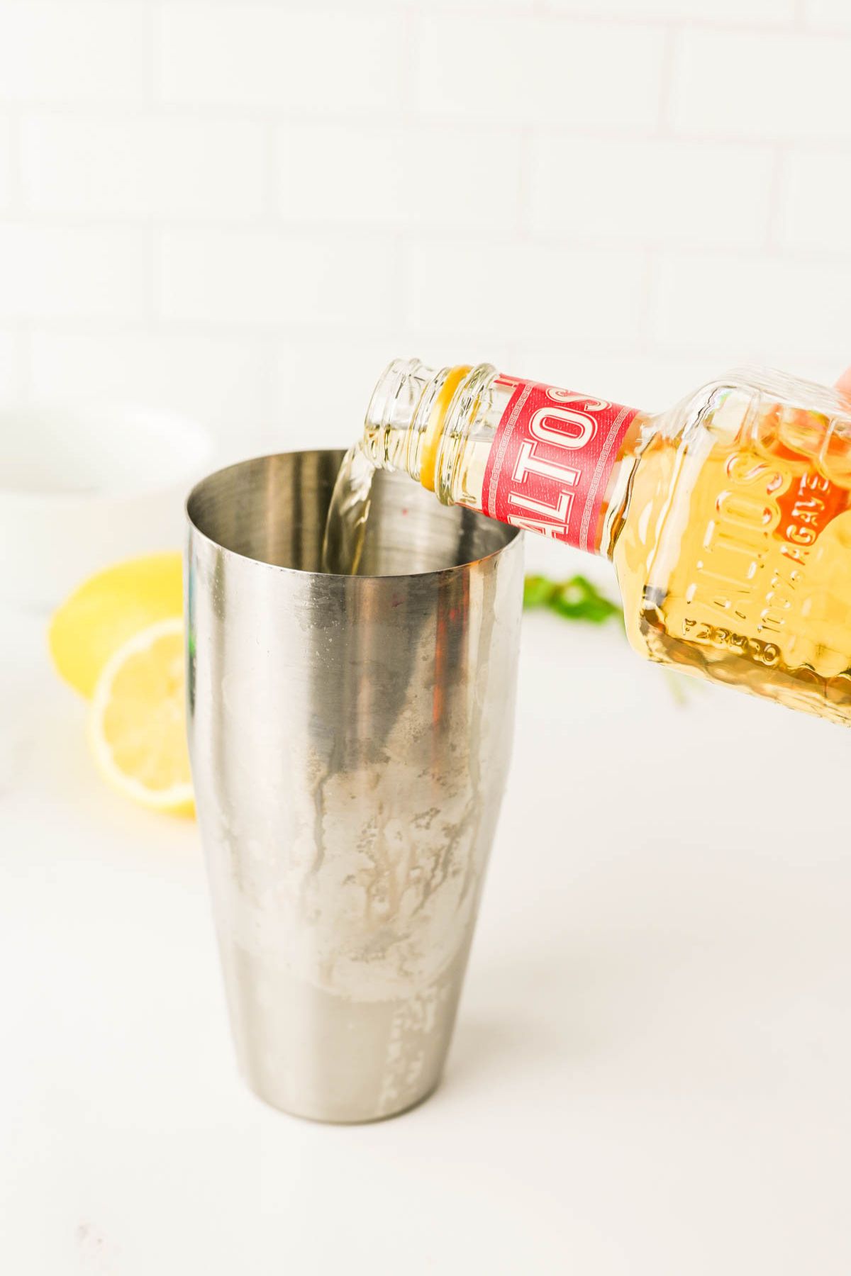 Stainless shaker with tequila pouring in from bottle and lemons in background.