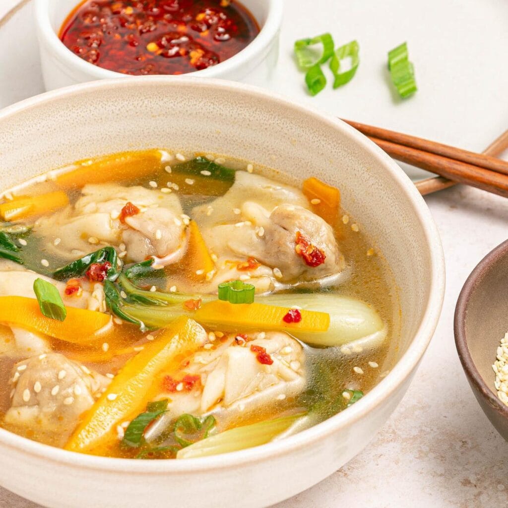 Bowl of chicken broth, vegetables, and wontons with sesame seeds and chili oil in a white bowl on a white counter with bowl of chili oil, bowl of sesame seeds, chopsticks, and sliced scallions scattered.