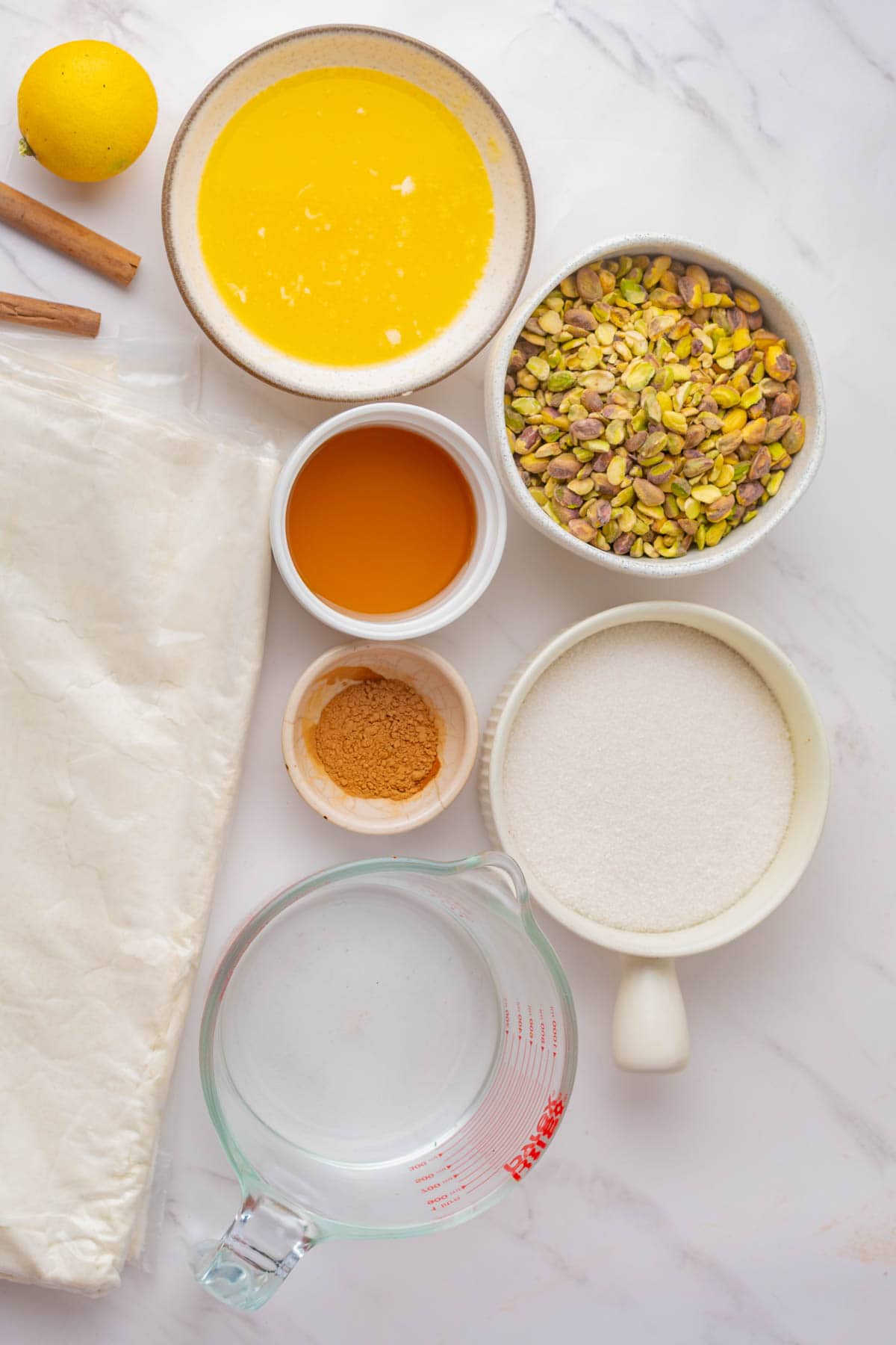Melted butter, lemon, cinnamon sticks, honey, phyllo dough sheets, ground cinnamon, sugar, and pistachio nuts on a white counter.