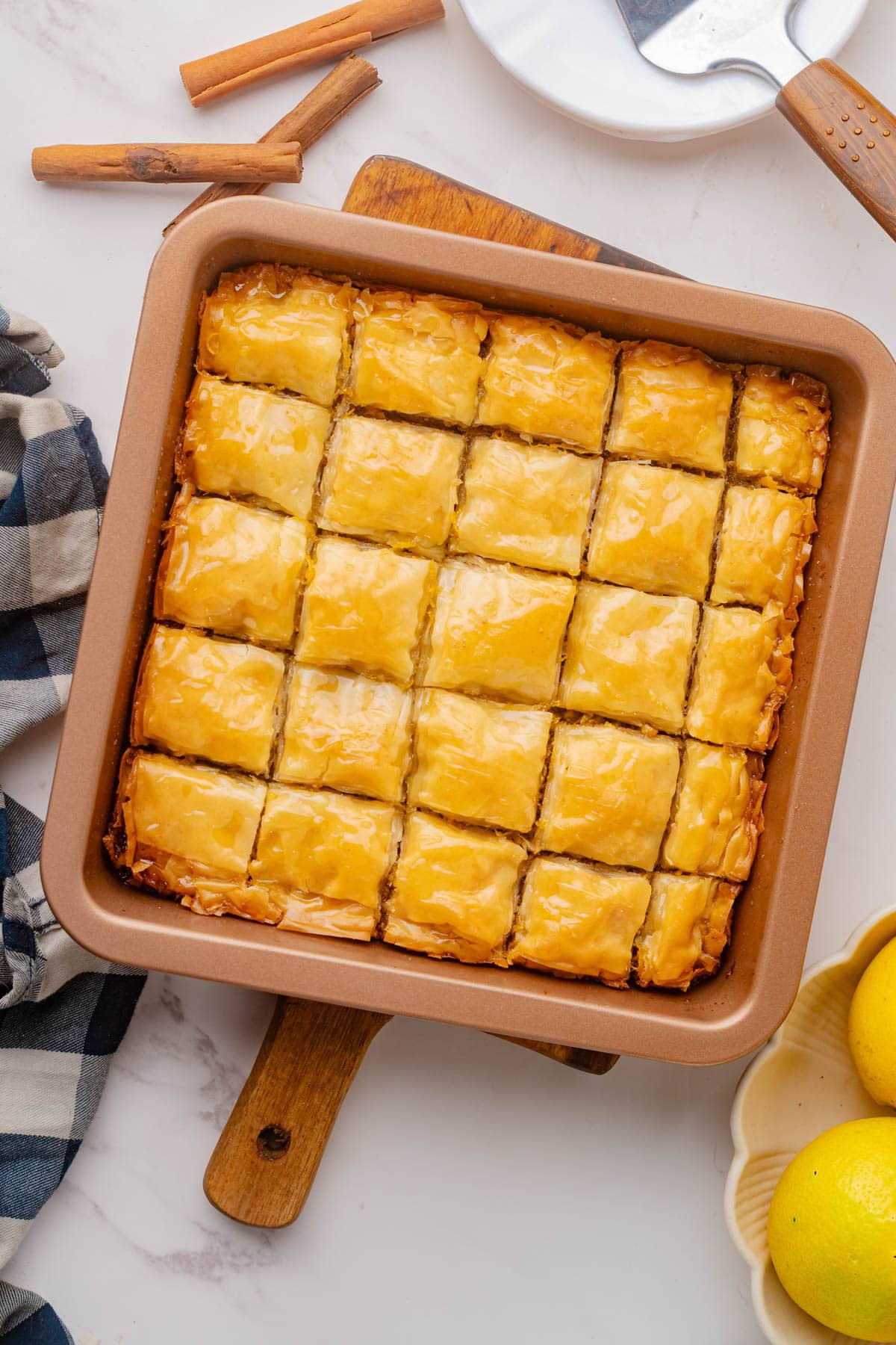 Baklava in a square baking pan on a wood board cut in small squares with bowl of lemons to the side and cinnamon sticks in above with a black and white dishtowel to the side..