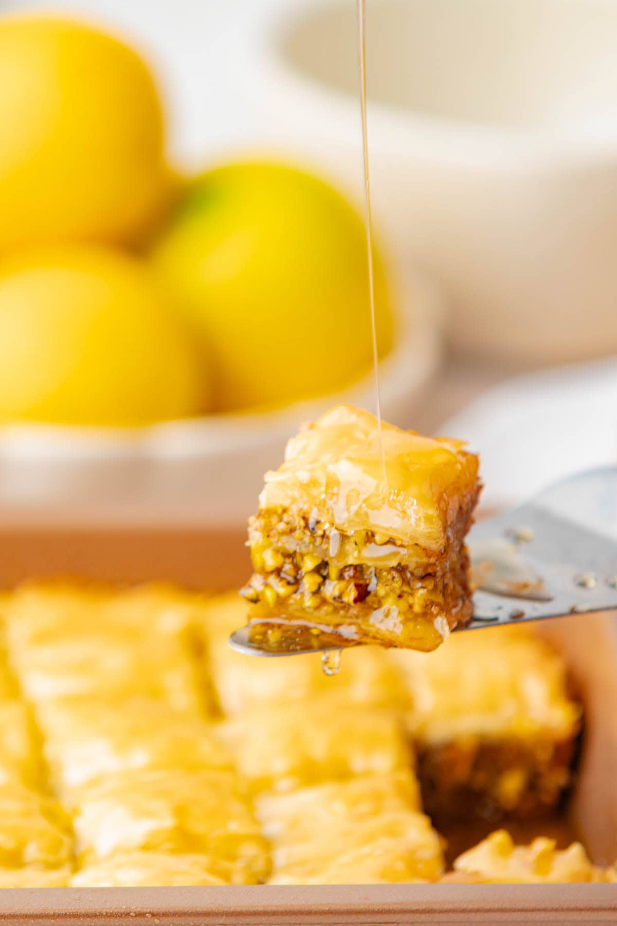 Piece of baklava on spatula with honey dripping from above and held above baking dish of more baklava.