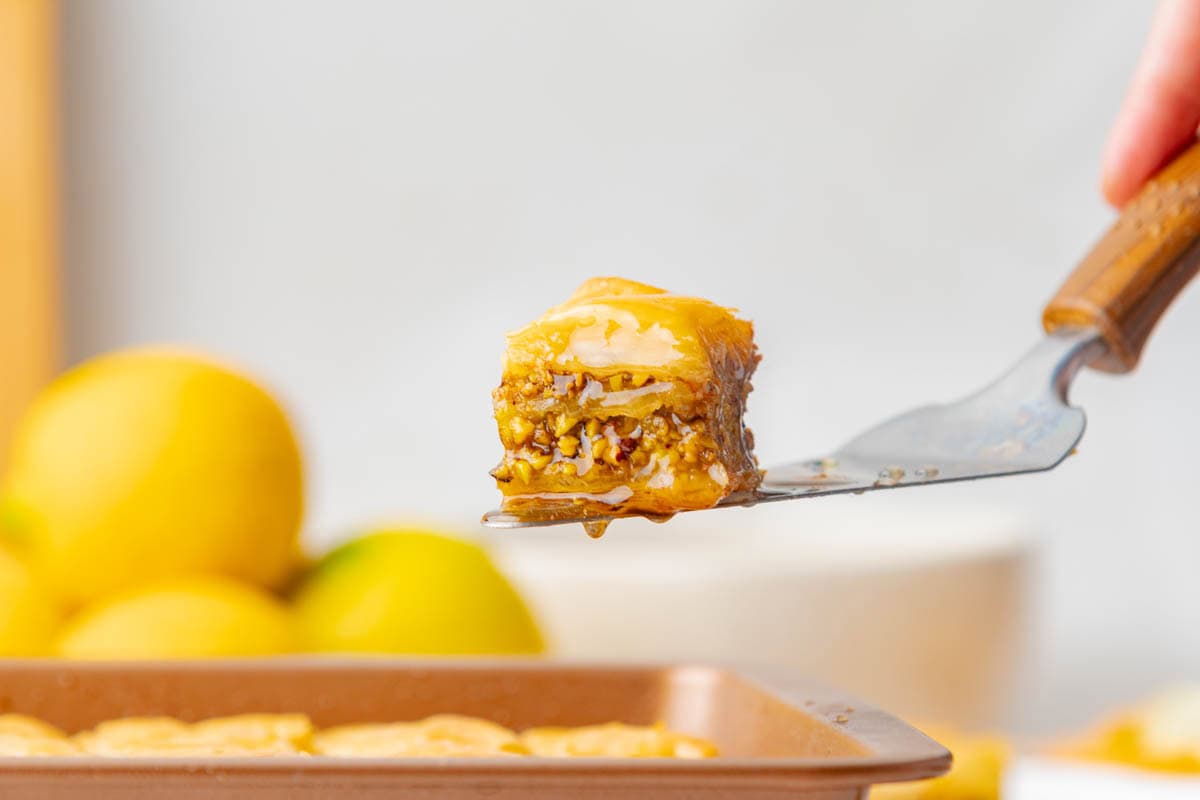 Piece of baklava on spatula with honey dripping at bottom and held above baking dish of more baklava.
