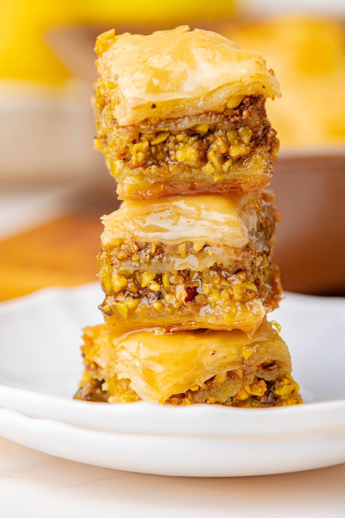 Stack of baklava pieces on a white plate with baking dish of baklava in background.
