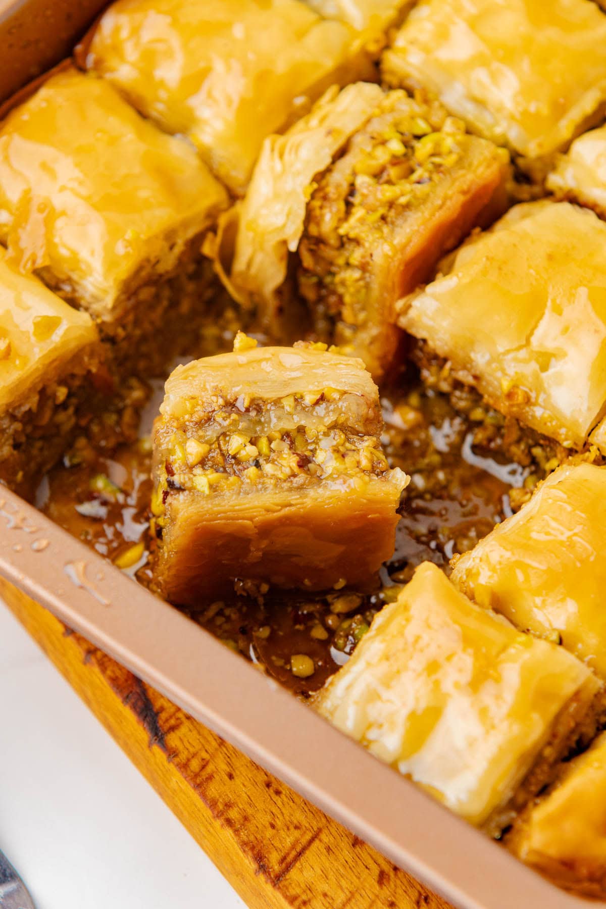 Baking dish of baklava cut into squares with pieces missing and one piece turned sideways laying in syrup at bottom.
