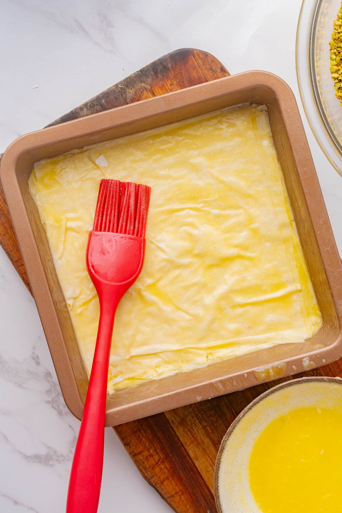 Phyllo dough in a square pan with a red brush and bowl of melted butter on a wood board.