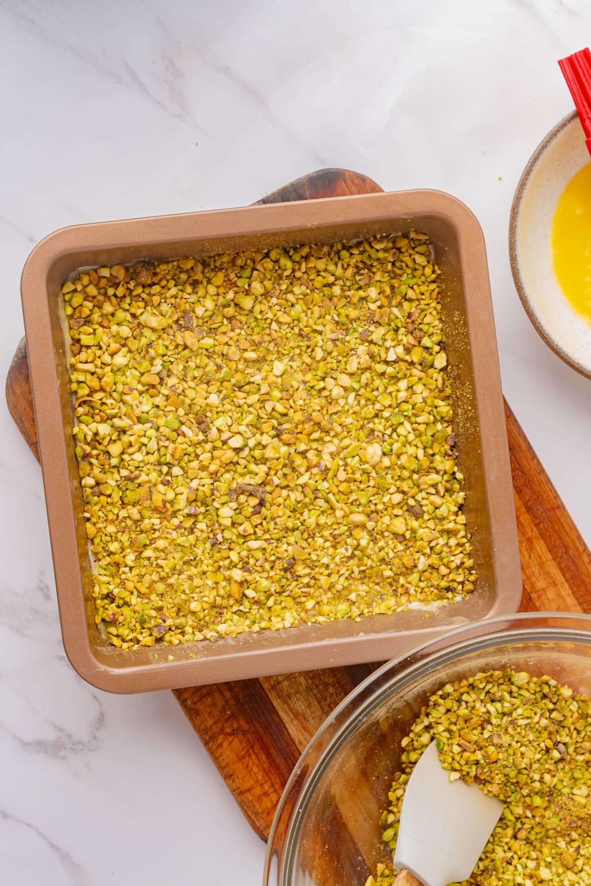 Phyllo dough covered in chopped pistachio nuts in a square pan and bowl of chopped pistachio nuts on a wood board with a bowl of melted butter to the side.