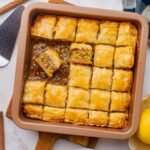 Baking dish of baklava cut into squares with pieces missing and one piece turned sideways laying in syrup at bottom.