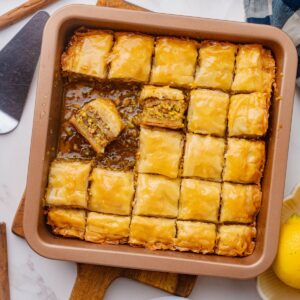Baking dish of baklava cut into squares with pieces missing and one piece turned sideways laying in syrup at bottom.