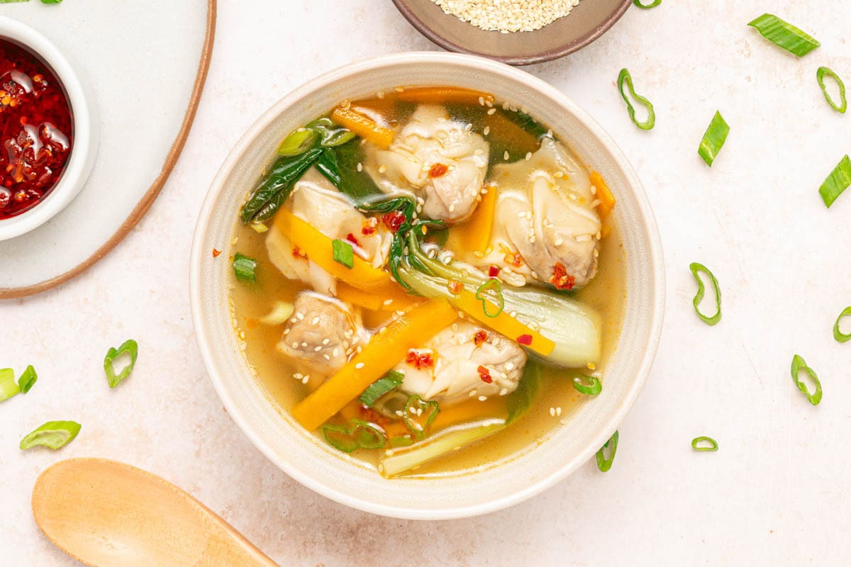Bowl of chicken broth, vegetables, and wontons with sesame seeds and chili oil in a white bowl on a white counter with bowl of chili oil, bowl of sesame seeds, wooden spoon, and sliced scallions scattered.