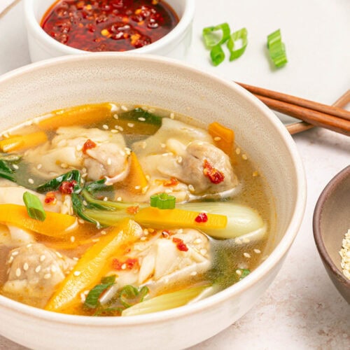 Bowl of chicken broth, vegetables, and wontons with sesame seeds and chili oil in a white bowl on a white counter with bowl of chili oil, bowl of sesame seeds, chopsticks, and sliced scallions scattered.