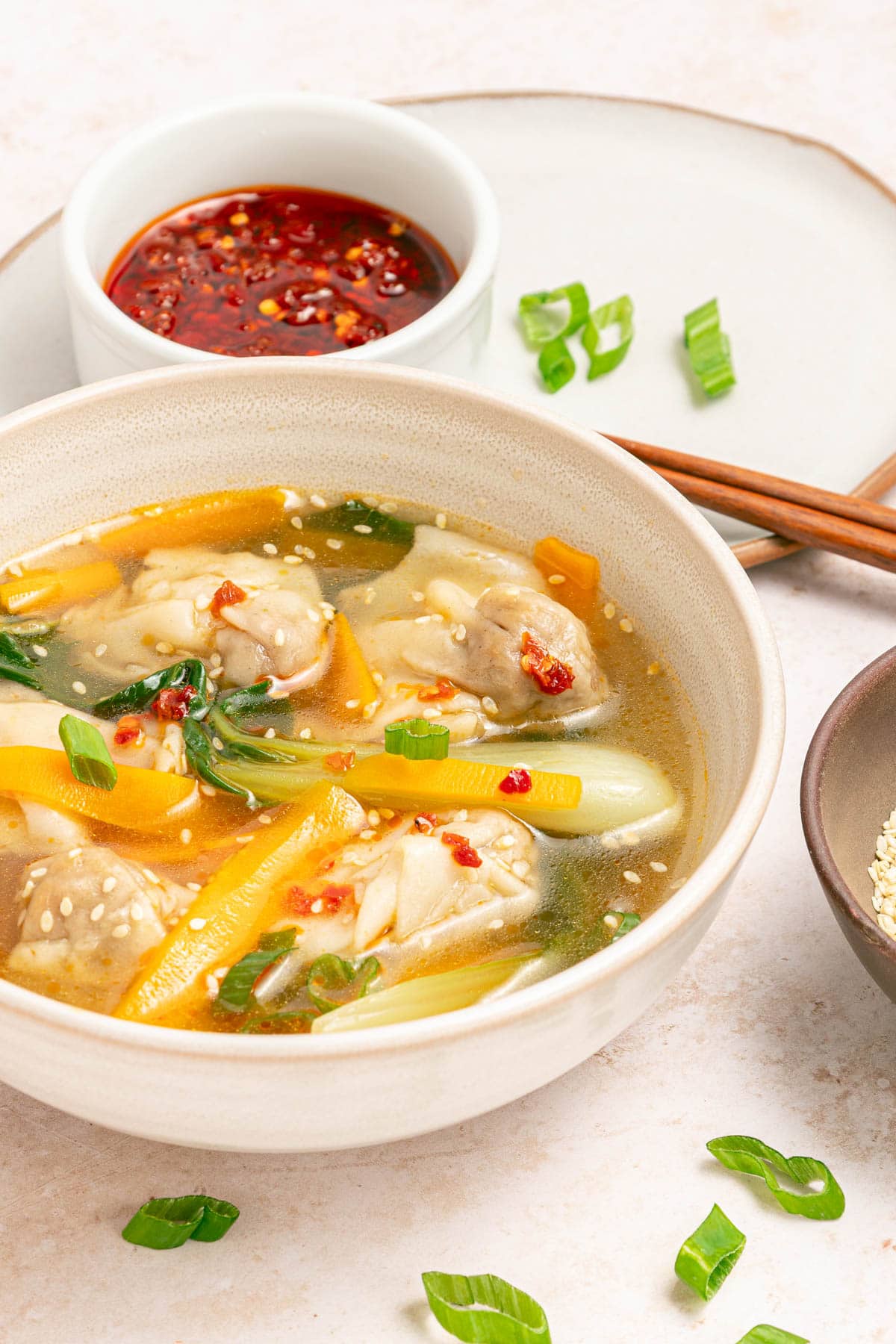 Bowl of chicken broth, vegetables, and wontons with sesame seeds and chili oil in a white bowl on a white counter with bowl of chili oil, bowl of sesame seeds, chopsticks, and sliced scallions scattered.