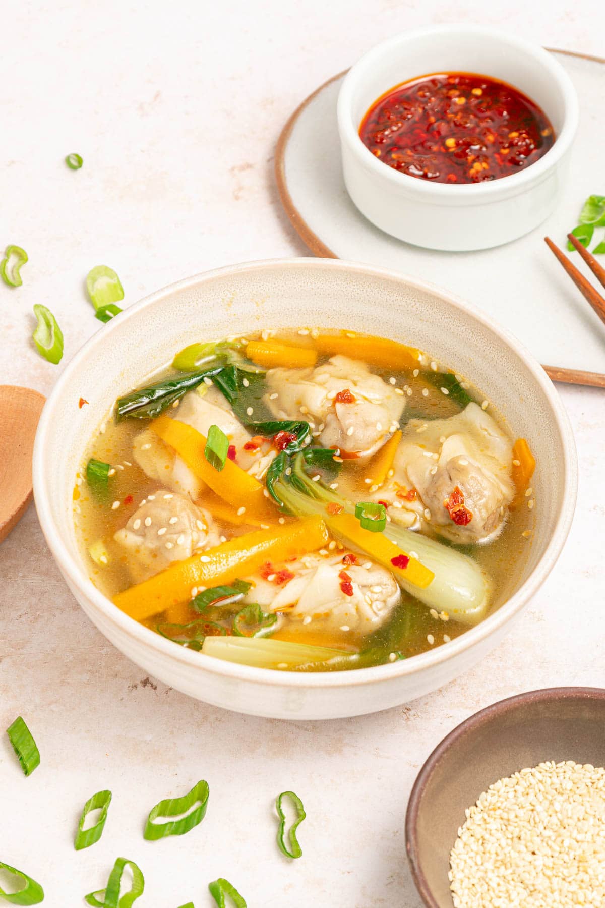 Chicken broth , vegetables, and wontons with sesame seeds and chili oil in a white bowl on a white counter with bowl of chili oil, bowl of sesame seeds, and sliced scallions scattered.