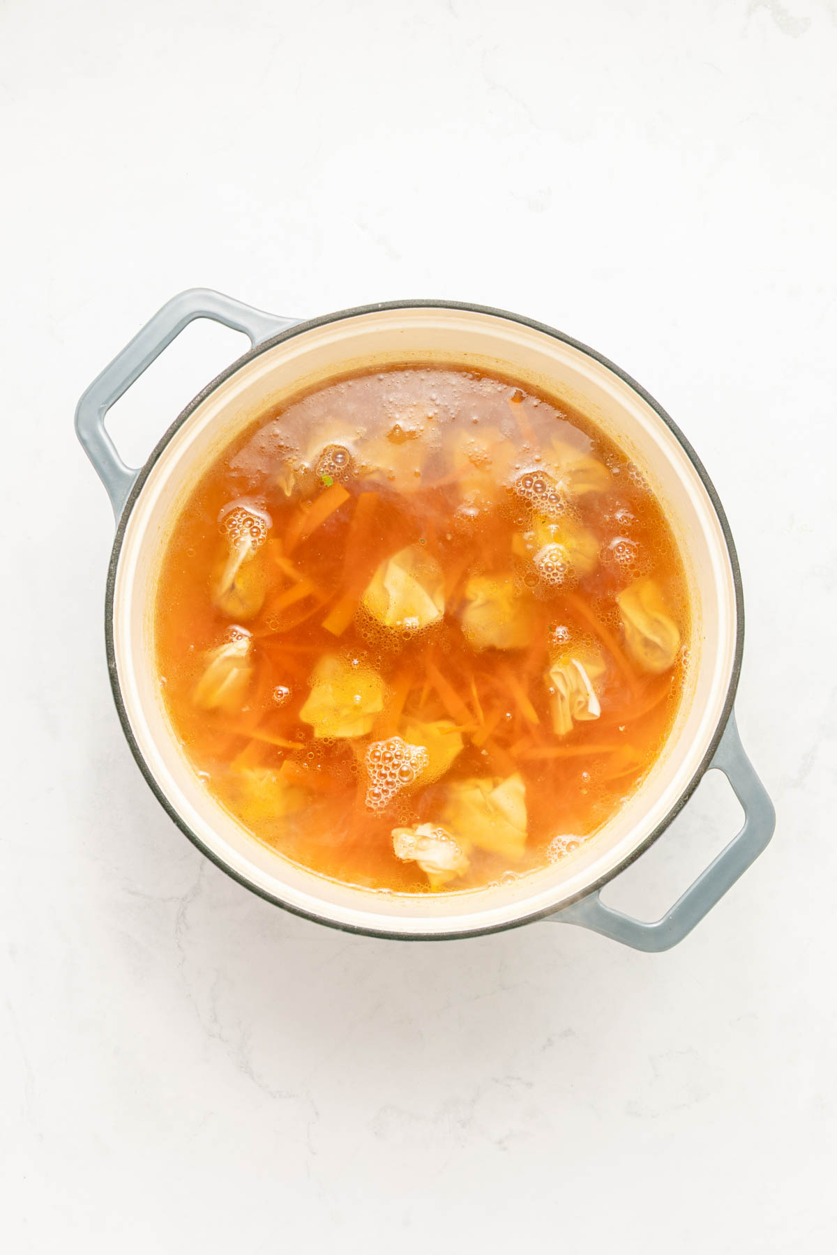 Chicken broth in a pot with carrots and wontons.