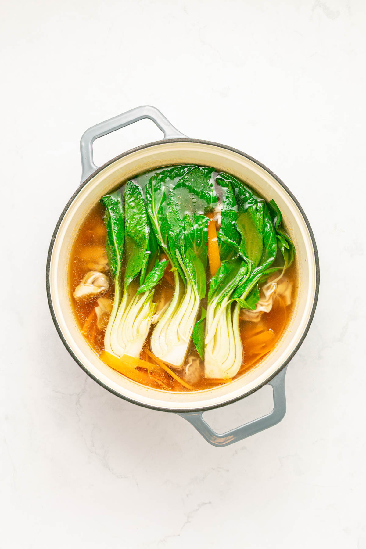 Chicken broth in a pot with carrots, wontons, and baby bok choy.