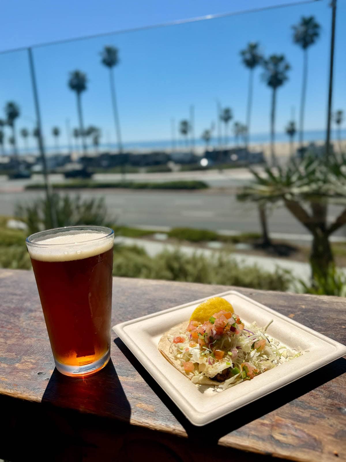 Fish taco on a white square plate with a beer set on a wood bar wih the beach in the background.