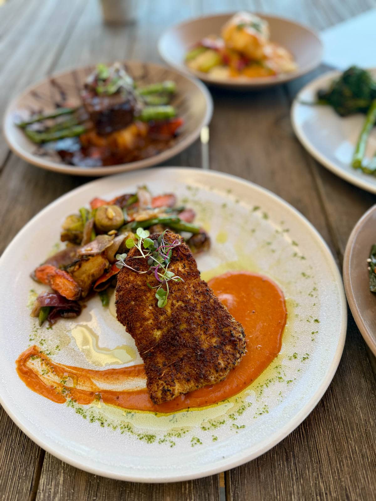 Panko- crusted fish on top of a red sauce with vegetables in the background on a white plate with other dishes behind.