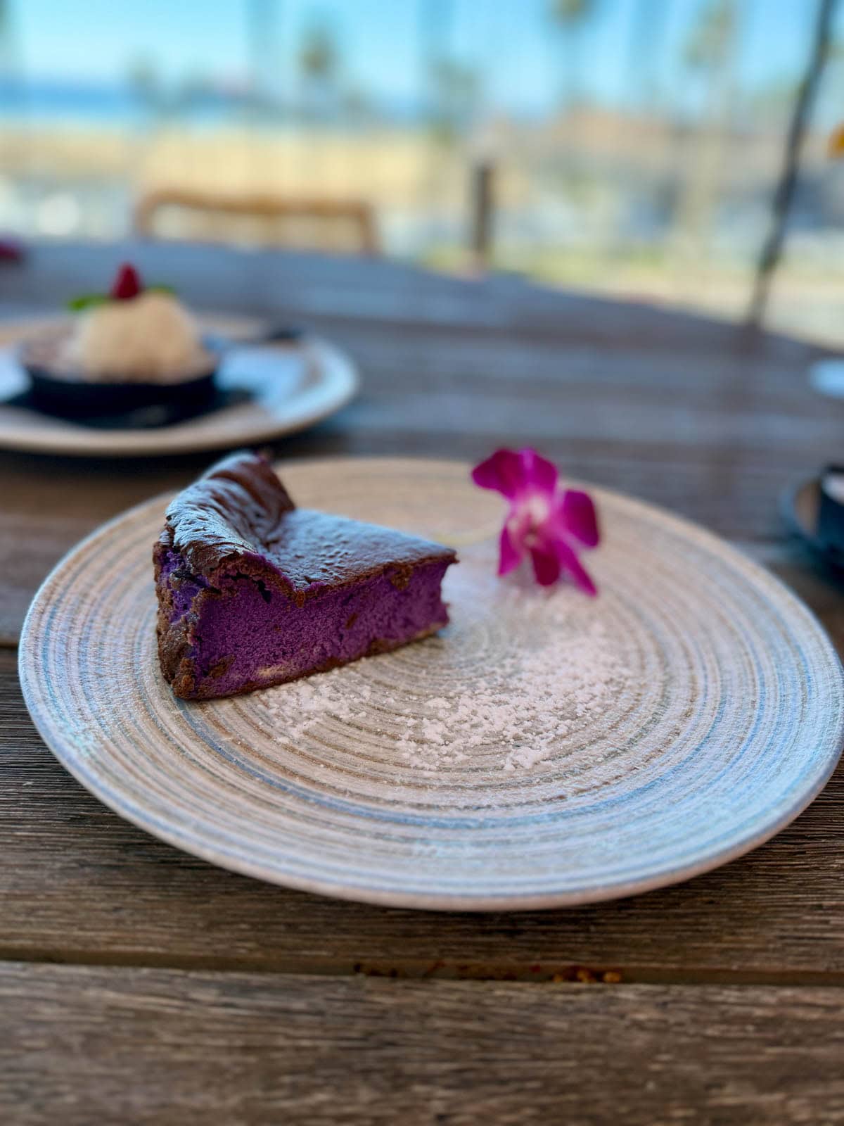 Slice of purple cake with powdered sugar and a purple flower on a grey plate.