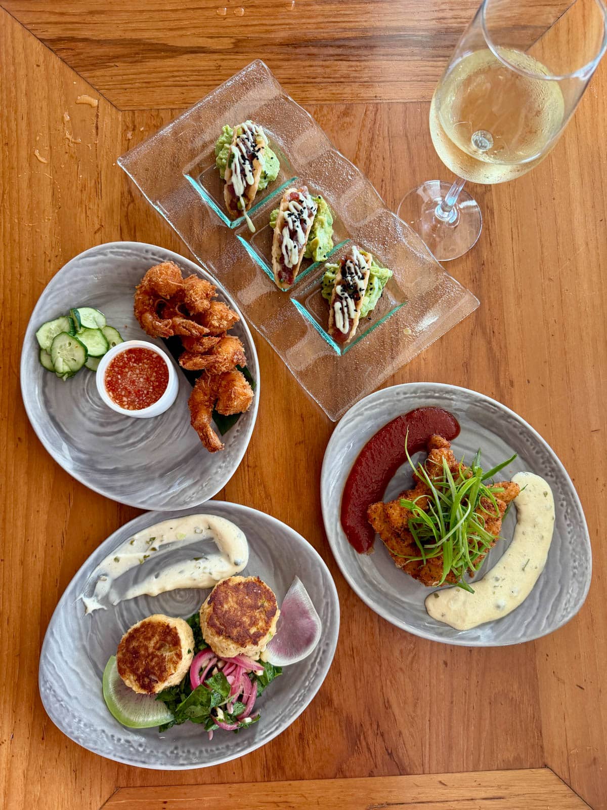 Tuna poke tacos, calamari tenders, crab cakes, and fried shrimp next to a glass of white wine on a wood table.