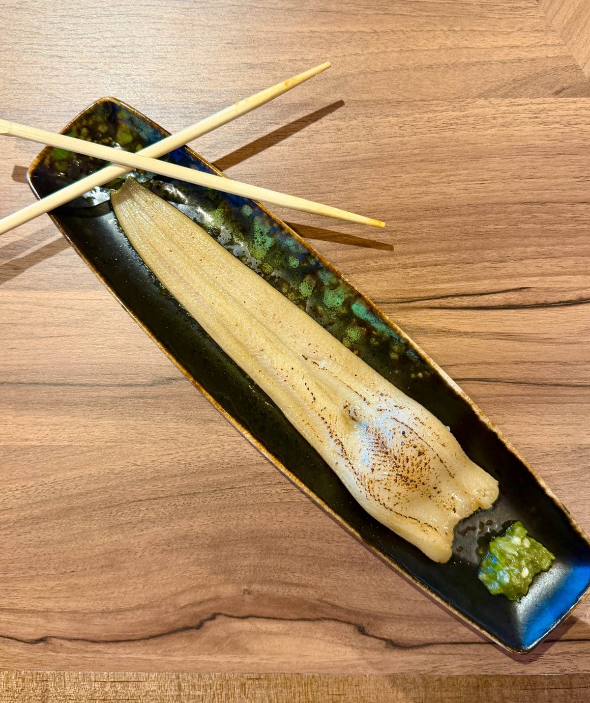 Long strip of seared eel on a long black plate with chopsticks and green sauce at one end.