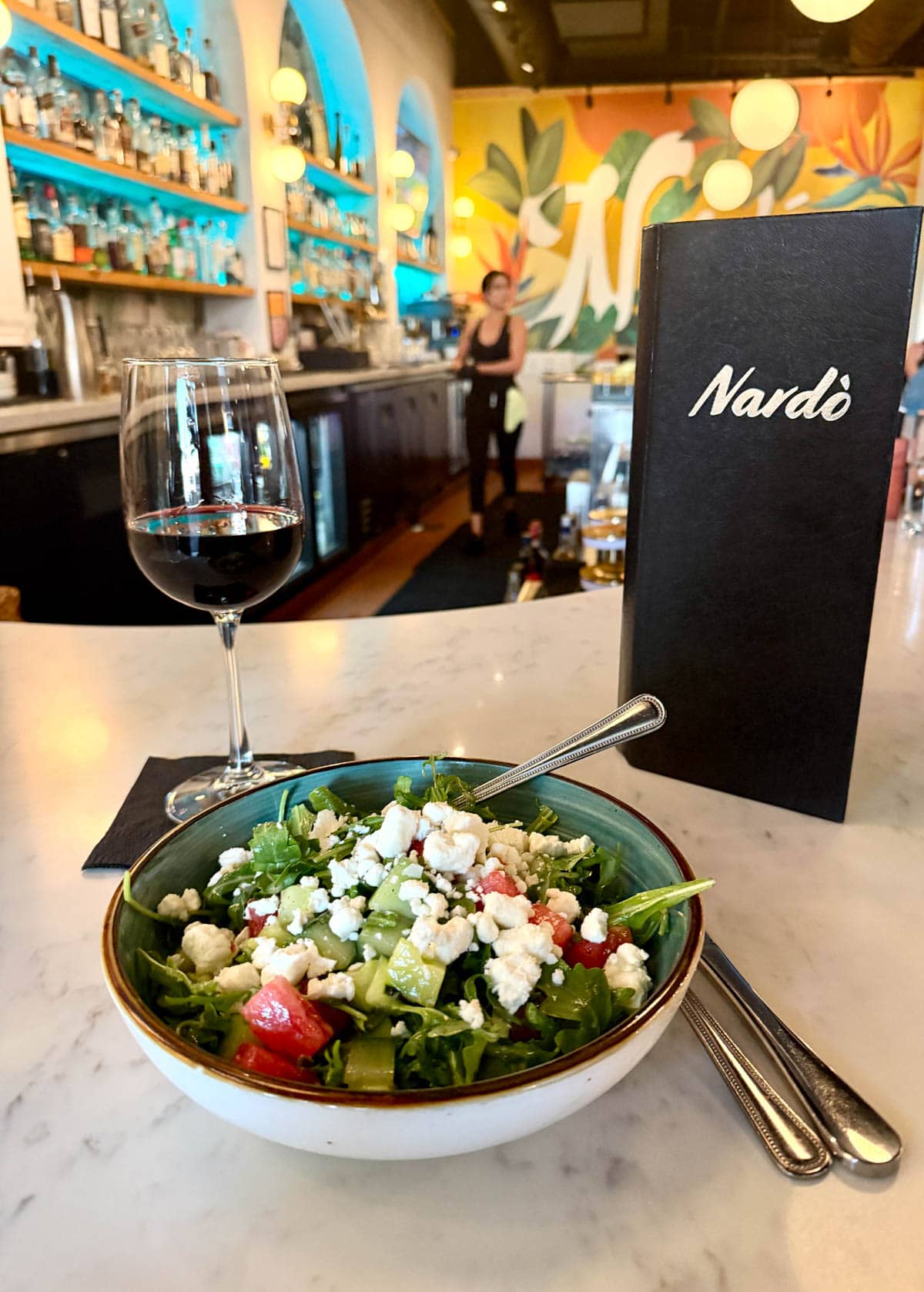 Watermelon salad with greens, cucumber, and feta cheese in a light grey bowl with a glass of wine and a menu in the background.