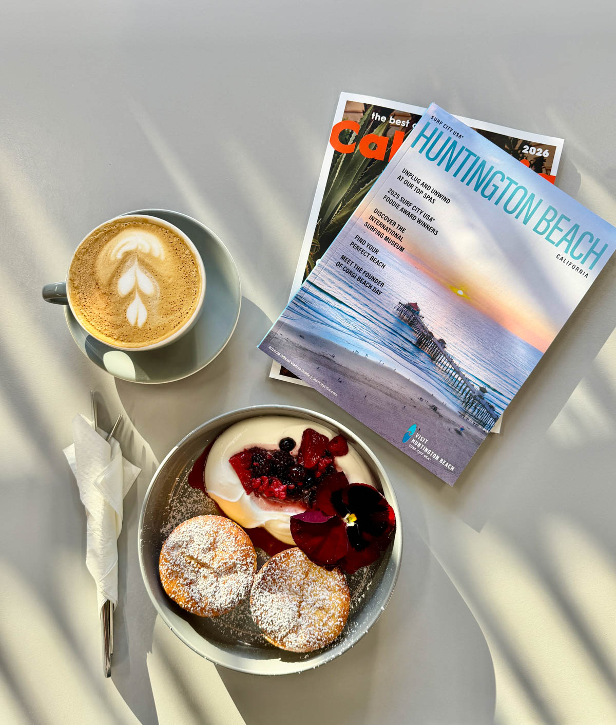 Pastries with yogurt and fruit in a grey bowl with a lattee coffee above on left and local magazines above on right.