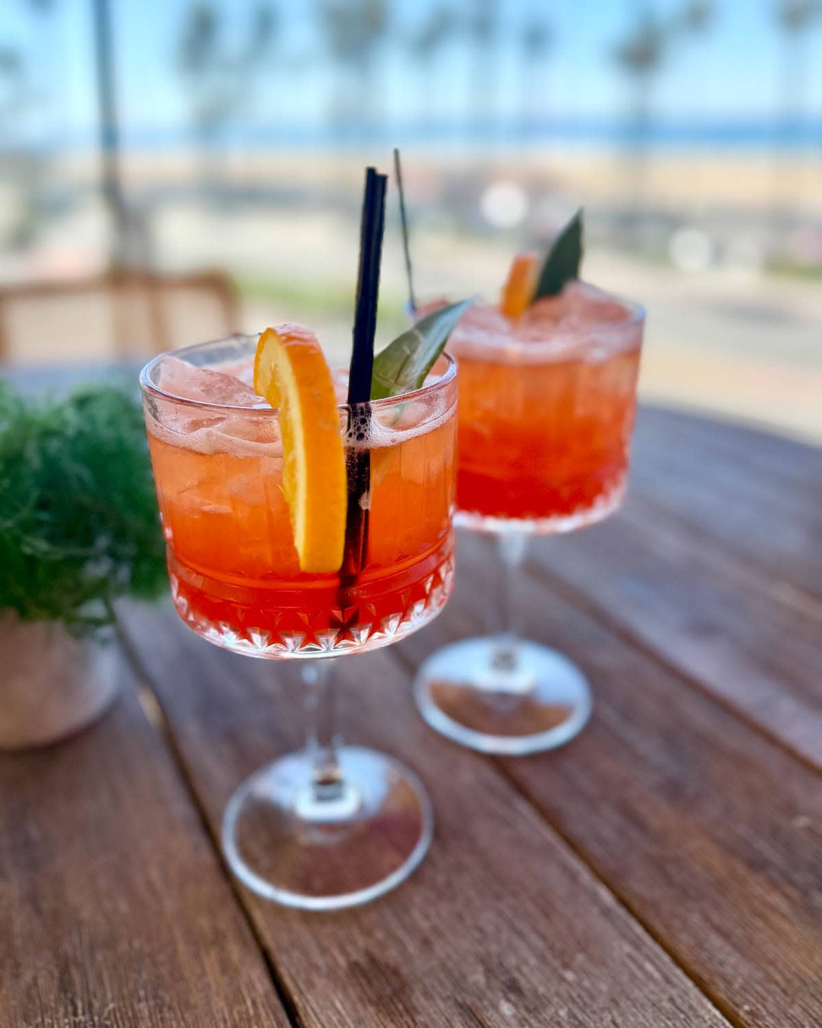 Two orange colored cocktails on a wood table with blurred view of beach in background