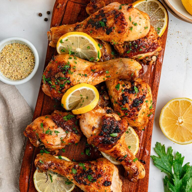 Cooked chicken drumsticks with lemon slices and fresh parsley on a wood cutting board with a handle, with parsley, lemon halves, and seasonings to the side.