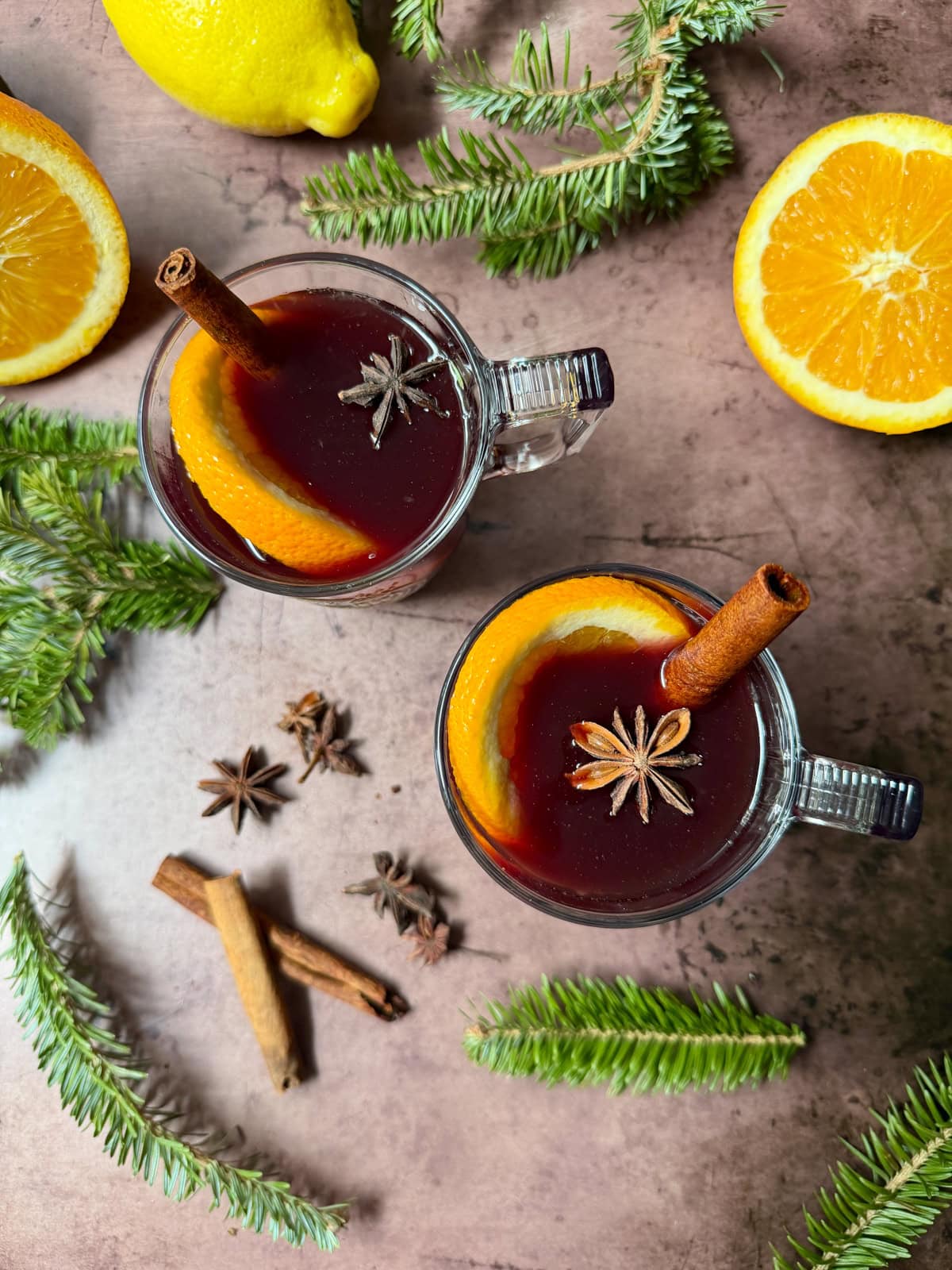 Glass mugs of red liquid with cinnamon stick, star anise, and orange slice, with more spices on the counter along with Christmas greens.