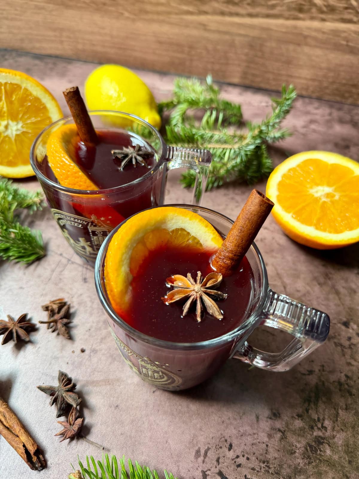 Glass mugs of red liquid with cinnamon stick, star anise, and orange slice, with more spices on the counter along with Christmas greens.