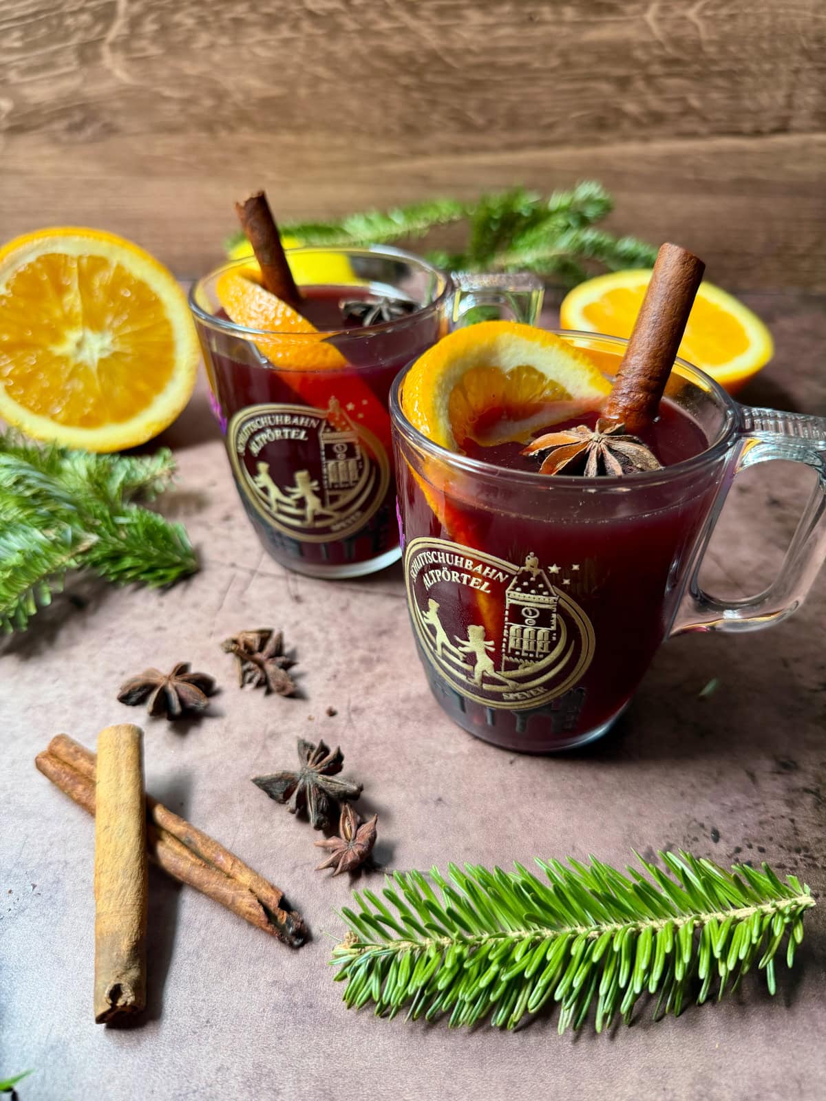 Glass mugs of red liquid with cinnamon stick, star anise, and orange slice, with more spices on the counter along with Christmas greens.