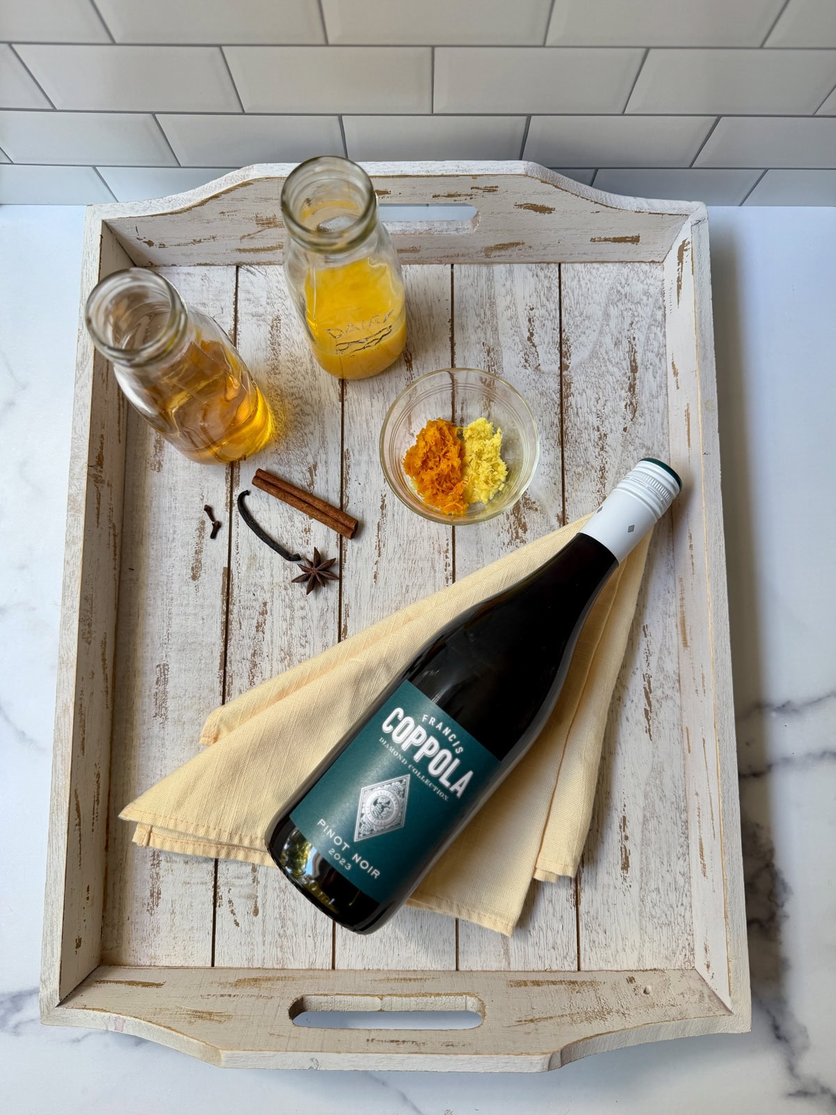 Bottle of wine, glass bowl of orange and lemon zest, cinnamon stick, vanilla bean, and star anise, and glass bottle of orange juice and glass bottle of apple juice.