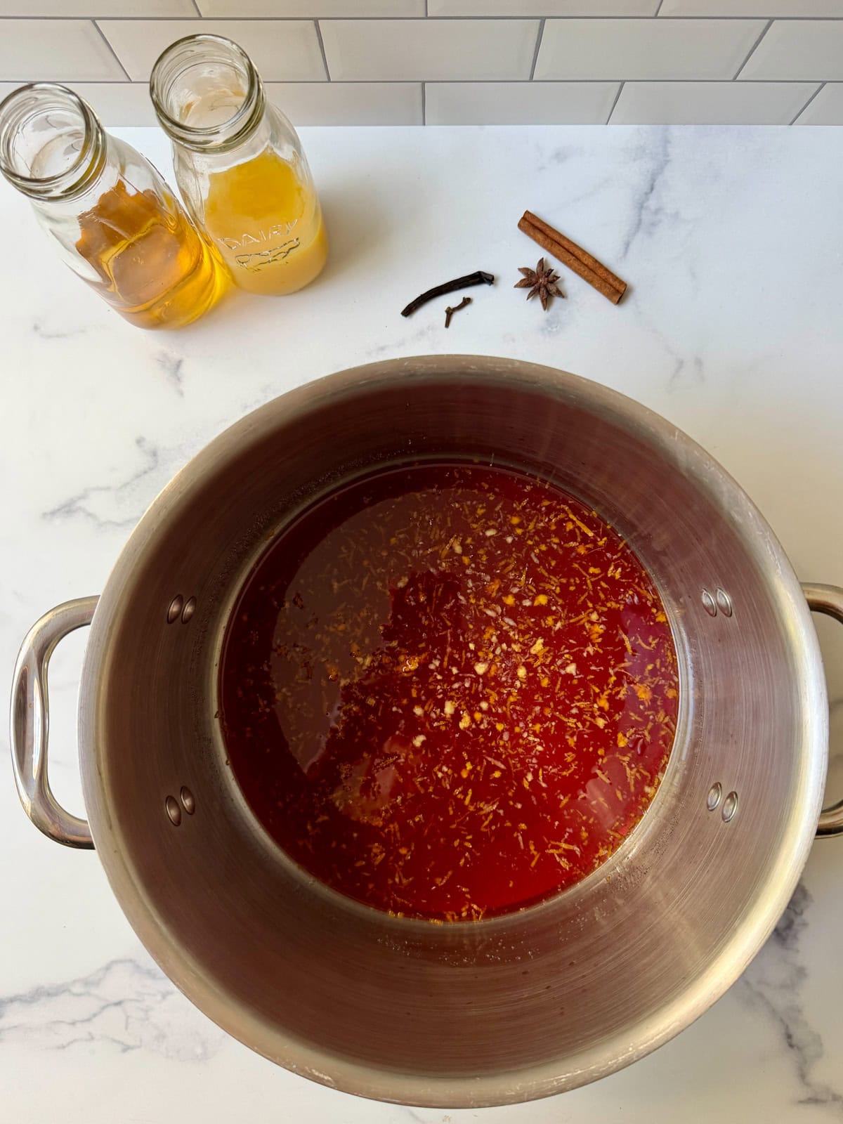 Stainless pot with dark red liquid and spices on a white counter with two glass jars and dried spices on white counter.