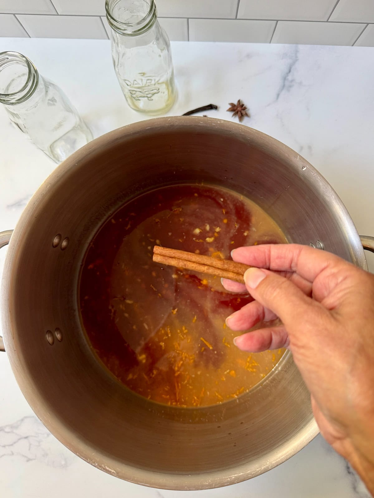 Stainless pot with dark red liquid and spices on a white counter with a hand holding a cinnamon stick above the pot.