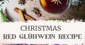 Glass mugs of red liquid with cinnamon stick, star anise, and orange slice, with more spices on the counter along with Christmas greens for Pinterest.