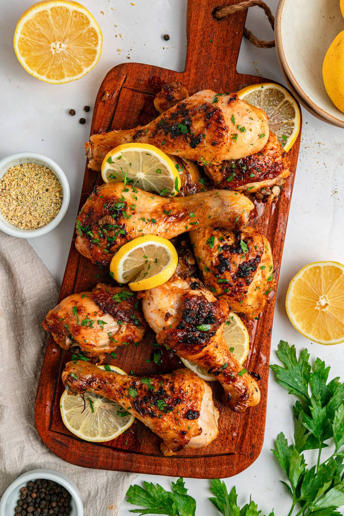 Cooked chicken drumsticks with lemon slices and fresh parsley on a wood cutting board with a handle, with parsley, lemon halved, and seasonings to the side.