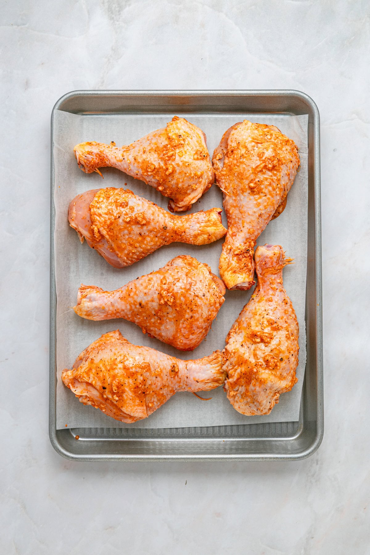 Marinade rubbed into chicken on parchment covered baking sheet on a white counter.